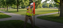 Person in high-visibility safety gear using a pole-mounted device in a park setting